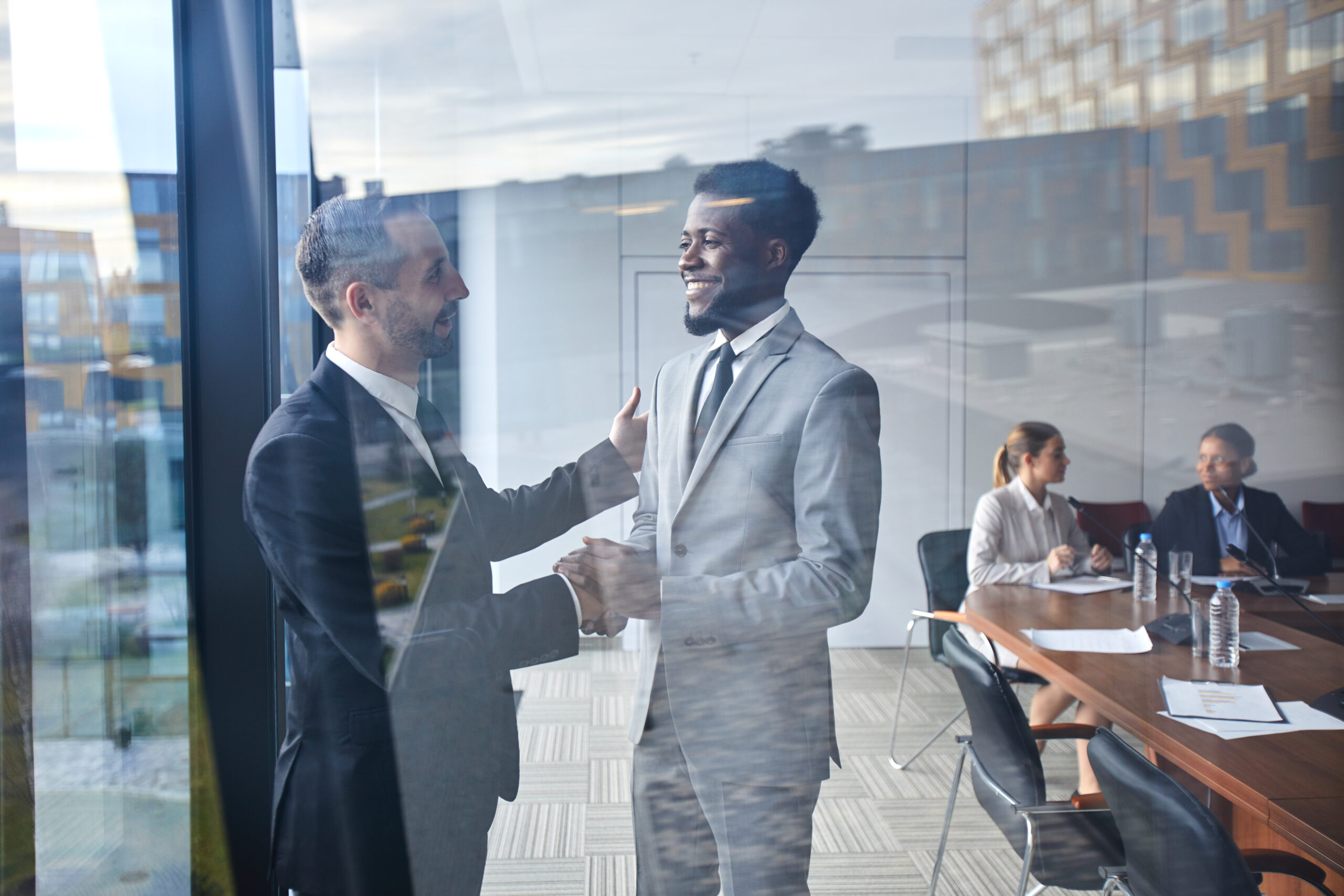 Two business professionals shaking hands in front of a glass wall after a successful meeting.